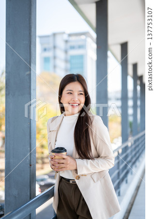 Pretty asian woman office worker holding coffee with both hands as standing or walking on footbridge Pretty asian woman office worker holding coffee with both hands as standing or walking on footbridge 137753107
