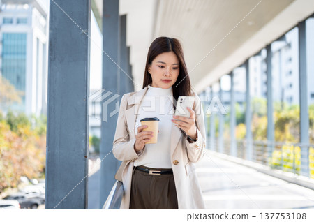 Pretty asian woman office worker holding coffee looking at phone standing or walking on a footbridge Pretty asian woman office worker holding coffee looking at phone standing or walking on a footbridge 137753108