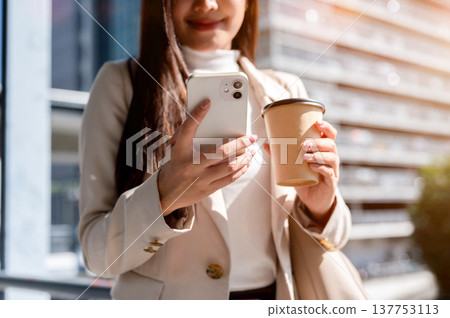 Close up of a woman in beige coat holding coffee cup looking at smartphone while standing or walking Close up of a woman in beige coat holding coffee cup looking at smartphone while standing or walking 137753113