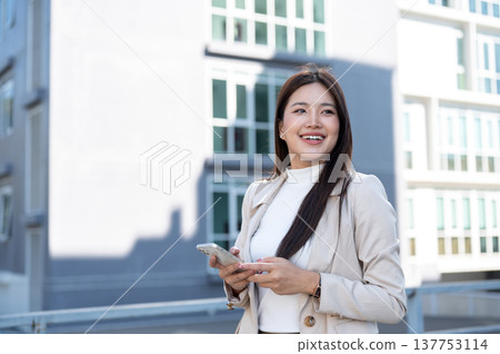 Pretty asian woman in beige coat holding phone looking away while standing or walking aside building 137753114