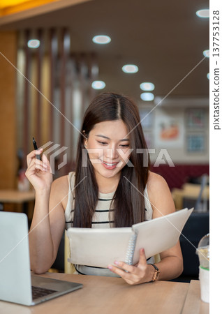 Pretty asian woman holding pen looking at notebook aside laptop and coffee sitting at table in cafe. Pretty asian woman holding pen looking at notebook aside laptop and coffee sitting at table in cafe. 137753128