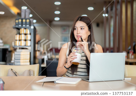 Pretty asian woman holding straw drinking coffee over laptop and notebook sitting at table in cafe. Pretty asian woman holding straw drinking coffee over laptop and notebook sitting at table in cafe. 137753137