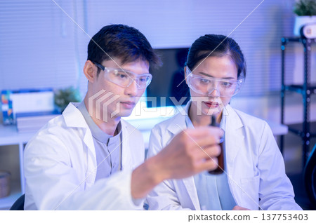 Scientist researcher man holding as looking at test liquid bottle with a woman colleague in lab room Scientist researcher man holding as looking at test liquid bottle with a woman colleague in lab room 137753403