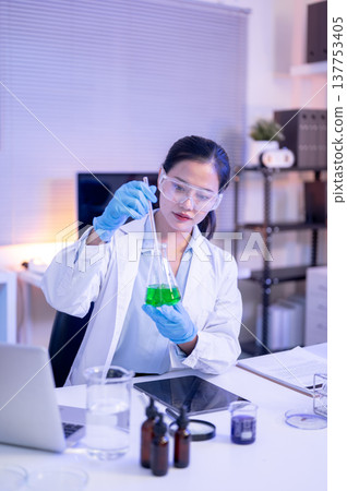 Woman scientist researcher wearing goggles holding glass flask testing liquid at table in lab room. 137753405