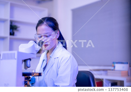 Asian woman scientist researcher using microscope looking at test sample in lab with purple lighting Asian woman scientist researcher using microscope looking at test sample in lab with purple lighting 137753408