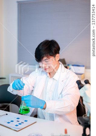 Scientist researcher man holding glass flask stirring test liquid over document in dim lit lab room. 137753414