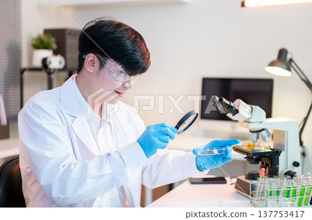 Scientist researcher man using magnifying glass look at test sample on dish aside microscope in lab. Scientist researcher man using magnifying glass look at test sample on dish aside microscope in lab. 137753417