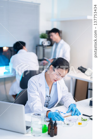 Woman scientist researcher examining test sample aside laptop while working with colleagues in lab. Woman scientist researcher examining test sample aside laptop while working with colleagues in lab. 137753434