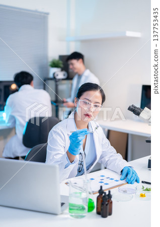 Woman scientist researcher looking at test sample aside laptop while working with colleagues in lab. Woman scientist researcher looking at test sample aside laptop while working with colleagues in lab. 137753435