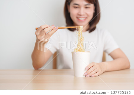Happy young Asian woman enjoying a delicious cup of hot instant noodles while relaxing at home, Cheerful female eating instant ramen with a smile at apartment. Simple lifestyle and quick meal concept Happy young Asian woman enjoying a delicious cup of hot instant noodles while relaxing at home, Cheerful female eating instant ramen with a smile at apartment. Simple lifestyle and quick meal concept 137755185