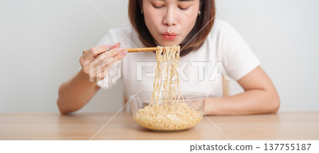 Happy young Asian woman enjoying a delicious bowl of hot instant noodles while relaxing at home, Cheerful female eating instant ramen with a smile at apartment. Simple lifestyle and quick meal concept Happy young Asian woman enjoying a delicious bowl of hot instant noodles while relaxing at home, Cheerful female eating instant ramen with a smile at apartment. Simple lifestyle and quick meal concept 137755187