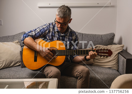 Man plays guitar on couch in living room during daytime Man plays guitar on couch in living room during daytime 137756256