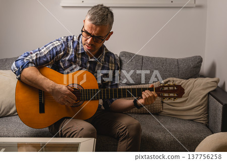 Man plays guitar while sitting on couch in modern living room Man plays guitar while sitting on couch in modern living room 137756258