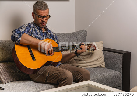 Man plays guitar while sitting on a couch in a living room Man plays guitar while sitting on a couch in a living room 137756270
