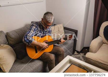Man plays guitar while sitting on couch in living room Man plays guitar while sitting on couch in living room 137756274