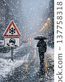 A person walks under snowfall with an umbrella on a snowy city street, illuminated by streetlights and icy road warning signs. 137758318