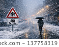 A person walks under snowfall with an umbrella on a snowy city street, illuminated by streetlights and icy road warning signs. 137758319