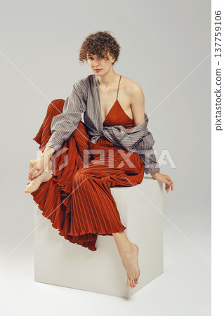 Stylish woman sits gracefully on a white cube against a neutral background Stylish woman sits gracefully on a white cube against a neutral background 137759106