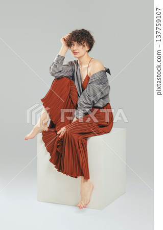 Elegant barefoot woman in stylish outfit sitting on studio cube 137759107