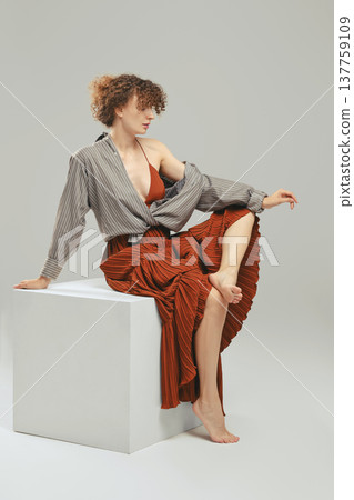 Barefoot woman in sundress and tied up shirt sits on wooden cube in the studio 137759109
