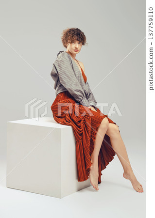 Young woman with curly hair in loose dress and shirt sits in profile on white cube against grey background Young woman with curly hair in loose dress and shirt sits in profile on white cube against grey background 137759110