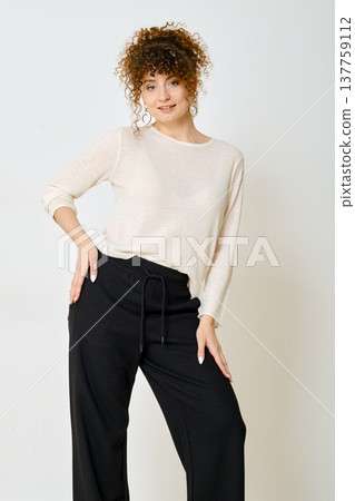 Woman with curly hair in white sweater and loose black pants leaned forward with her hand on her hip 137759112