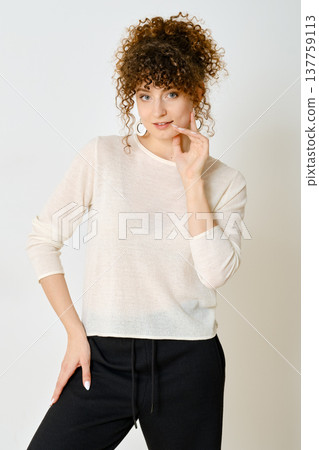 Curly-haired model posing in a light pullover and black pants against white wall 137759113