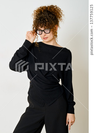 Stylish woman with curly hair wearing black outfit and glasses poses confidently in studio setting Stylish woman with curly hair wearing black outfit and glasses poses confidently in studio setting 137759125