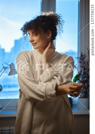 Young woman stretches her neck by the window in an urban setting during the late afternoon 137759135