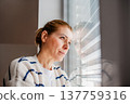 Beautiful woman looking through window at home. 137759316