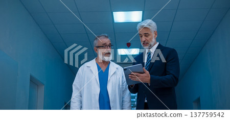 Manager walking with doctor down the hallway, showing him document in tablet. Manager walking with doctor down the hallway, showing him document in tablet. 137759542