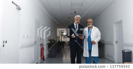 Manager walking with doctor down the hallway, showing him document in tablet. Manager walking with doctor down the hallway, showing him document in tablet. 137759543
