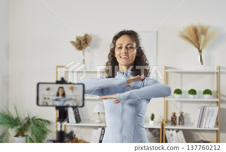 Smiling young woman recording fitness video on smartphone and showing warm up exercise at home. 137760212