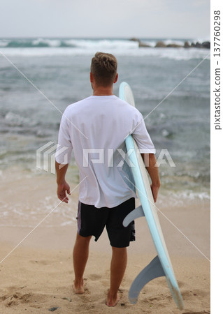 Relaxed Man With Surfboard At Shoreline. Man In White Tshirt Holding Surfboard On Wet Sand Relaxed Man With Surfboard At Shoreline. Man In White Tshirt Holding Surfboard On Wet Sand 137760298