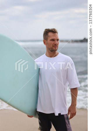 Beach Surfer Portrait. Closeup Image Of Surfer On Shoreline Sporting Relaxed Outfit Under Cloudy Sky 137760304