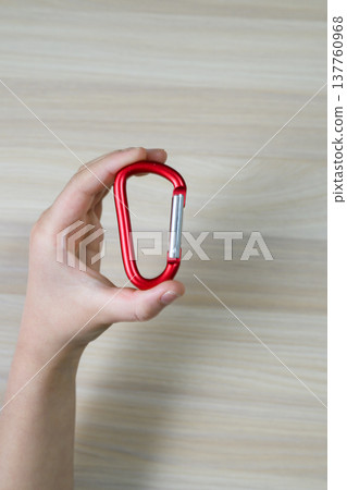 A hand holding a red carabiner against a wood-grain background (vertical). 137760968