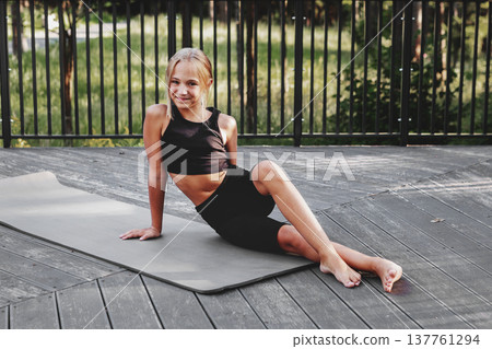 Preteen girl sitting on yoga mat in sportswear outdoors in forest after fitness training, smiling and looking at camera. Healthy child lifestyle, active recreation in nature, Athletic kid 137761294
