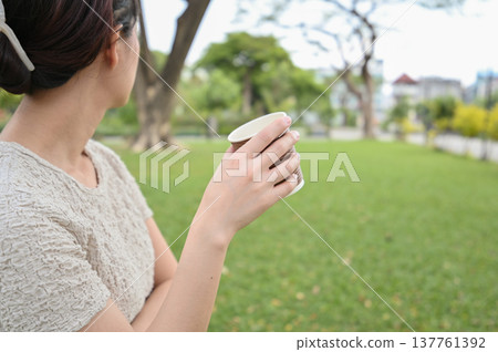 Contemplative asian woman holding paper cup of coffee relaxing in green park. peaceful moment outdoors with beverage, enjoying beautiful day in garden 137761392