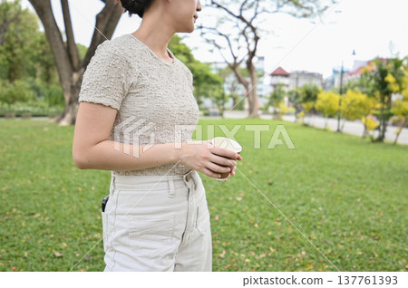 Thoughtful young woman standing outdoors in park. Relaxed female enjoying quiet moment with cup of coffee on green grass. peaceful, casual lifestyle concept 137761393