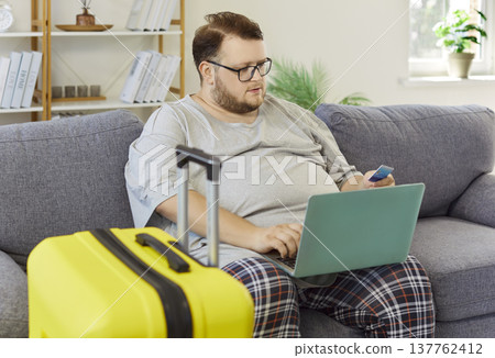 Smiling man paying for vacation on laptop at home with yellow suitcase Smiling man paying for vacation on laptop at home with yellow suitcase 137762412