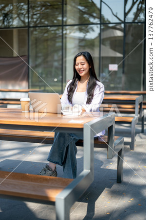 Pretty asian woman smiling as looking at laptop aside headphone sitting at outdoor table in sunlight 137762479