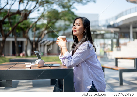 Pretty asian woman holding coffee cup over laptop and headphone sits at outdoor table under sunlight 137762480