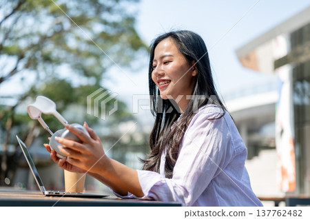 Smiling asian woman holding headphones aside laptop and coffee sitting at outdoor table in sunlight. Smiling asian woman holding headphones aside laptop and coffee sitting at outdoor table in sunlight. 137762482