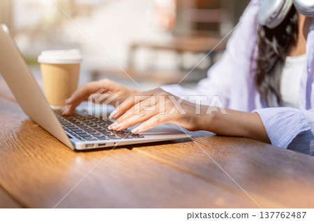 Close up of woman's hands typing on laptop aside coffee while sitting at wooden table under sunlight Close up of woman's hands typing on laptop aside coffee while sitting at wooden table under sunlight 137762487