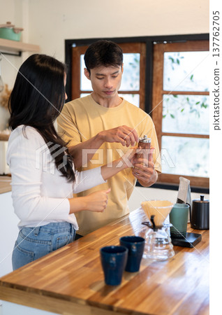 Asian couple a man holding grinder teaching a girlfriend making coffee at wooden counter in kitchen. Asian couple a man holding grinder teaching a girlfriend making coffee at wooden counter in kitchen. 137762705