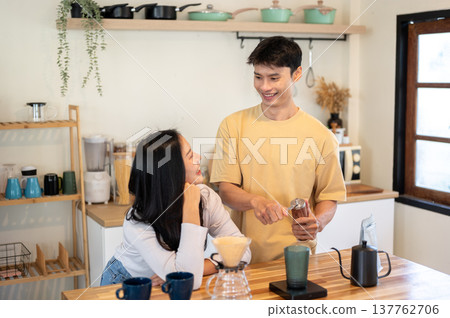 Asian man and woman couple looking talking together while making coffee at wooden counter in kitchen Asian man and woman couple looking talking together while making coffee at wooden counter in kitchen 137762706