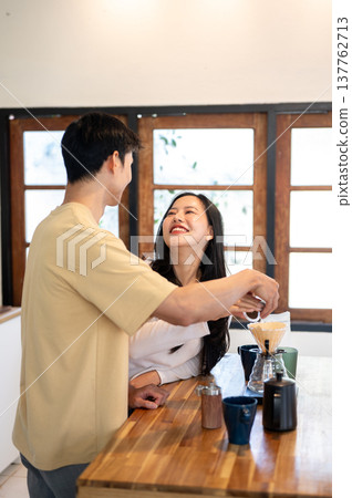 Asian man holding smiling woman's hand pouring powder in dripper making coffee. Couple in kitchen. Asian man holding smiling woman's hand pouring powder in dripper making coffee. Couple in kitchen. 137762713