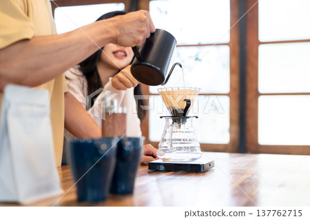 Close up of hand holding kettle pouring water in dripper making coffee with woman at kitchen counter Close up of hand holding kettle pouring water in dripper making coffee with woman at kitchen counter 137762715