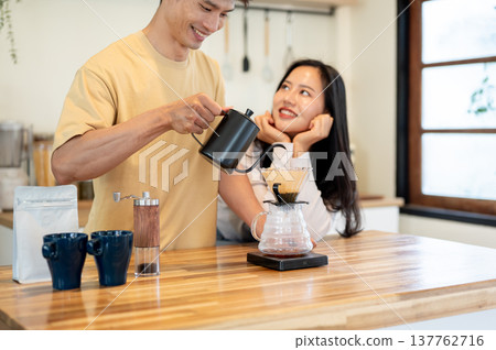 Asian woman looking at man holding kettle pouring water in dripper making coffee at kitchen counter. 137762716
