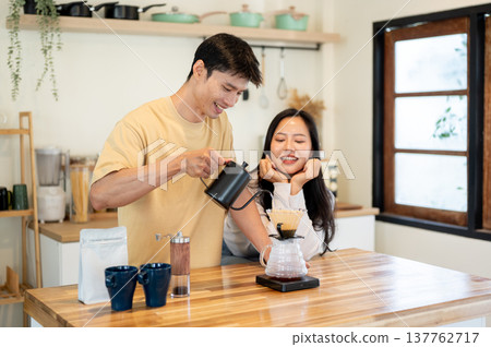 Asian woman looking at man holding kettle pouring water in dripper making coffee on kitchen counter. Asian woman looking at man holding kettle pouring water in dripper making coffee on kitchen counter. 137762717